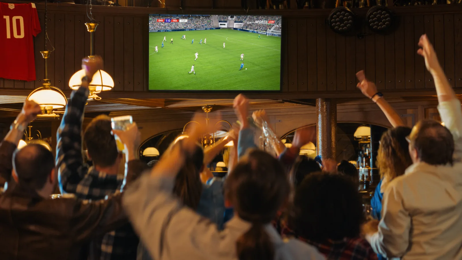 a crowd seen from behind with arms raised, watching a football match on a wall-mounted screen in a wood-paneled bar with brass pendant lamps