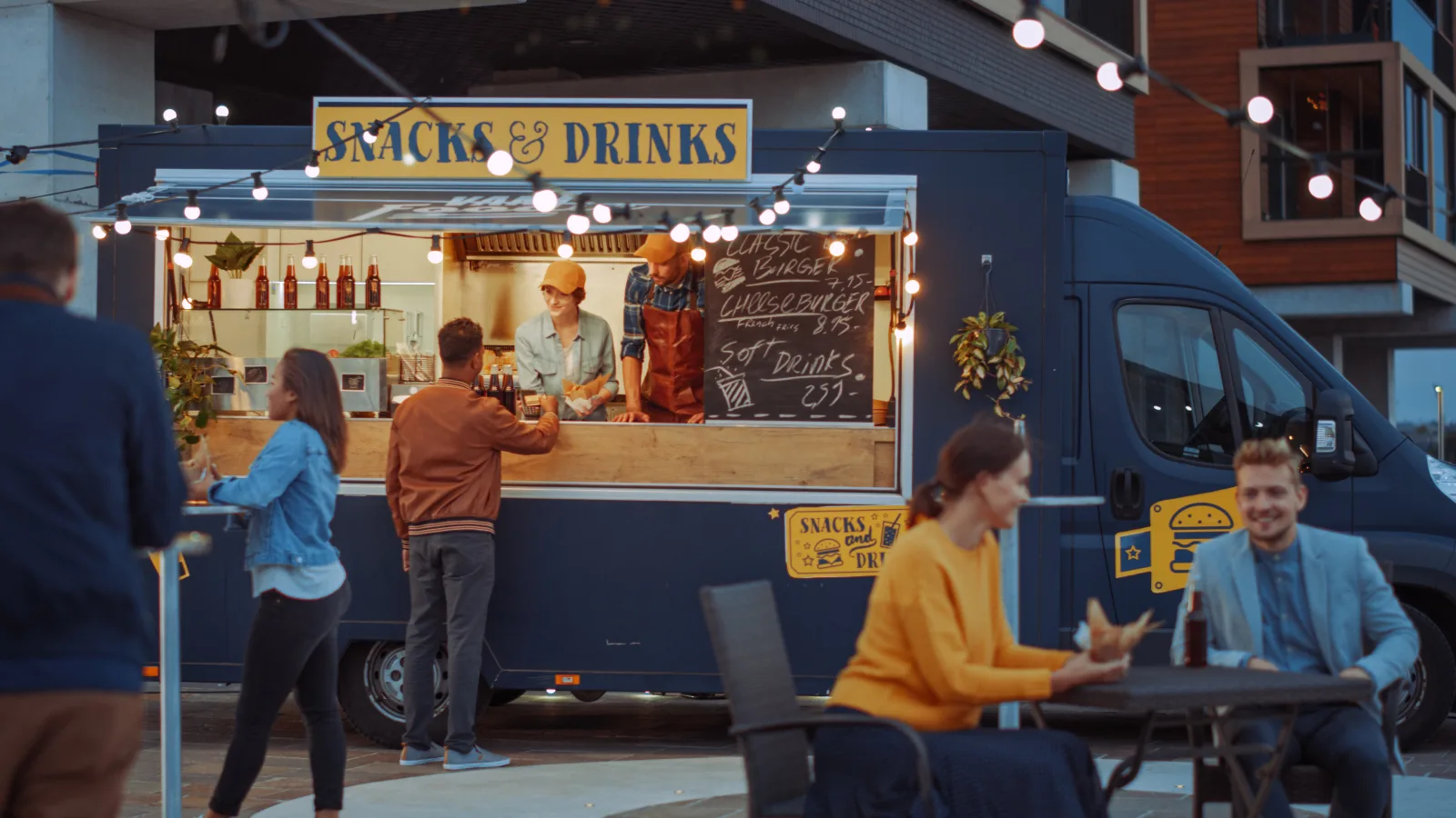 a dark blue food truck with a lit service window and a hand-lettered chalkboard menu, customers waiting at the hatch under strings of bulbs at dusk — general liability for food and drink operators
