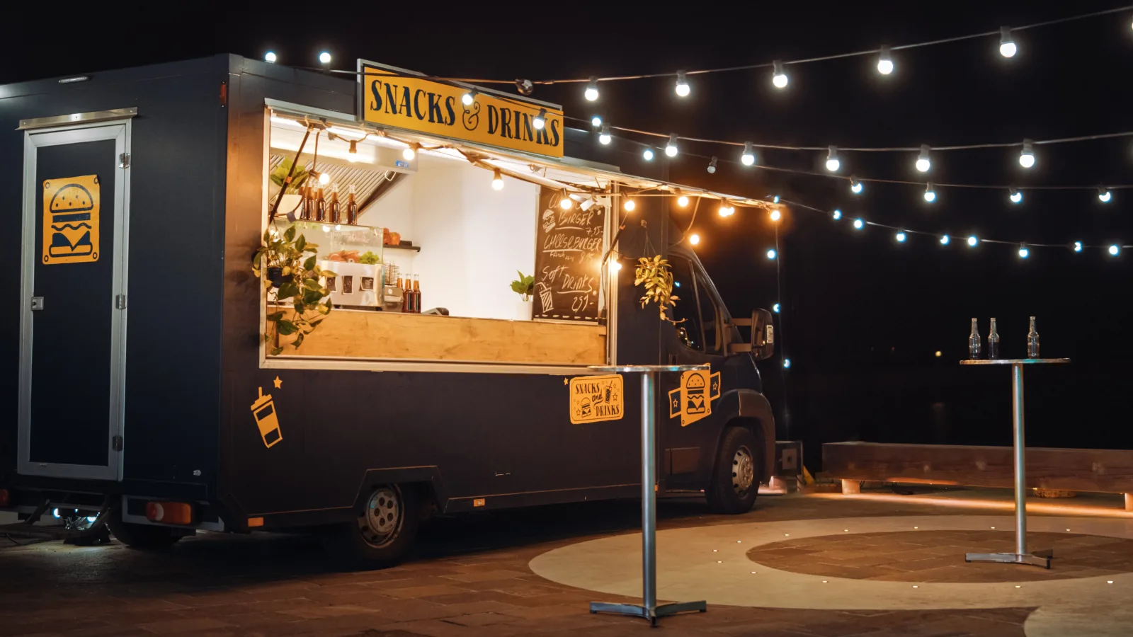 a dark blue food truck parked alone at night, its service window and awning lit, with strings of bulbs overhead and standing tables nearby — workers compensation for kitchen and floor staff
