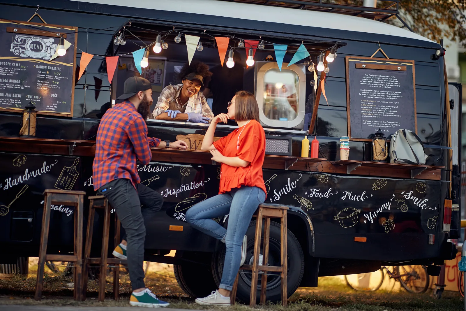 a converted black bus fitted out as a food truck, chalkboard menus and bunting along its flank, two customers on stools at the counter ledge — the operator types this brand insures