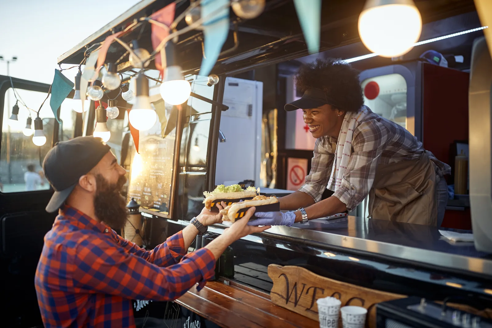 a customer taking a filled roll from a staff member leaning out of a service hatch, string lights and a fridge visible inside, low sun behind — starting a quote for a restaurant, bar or food truck