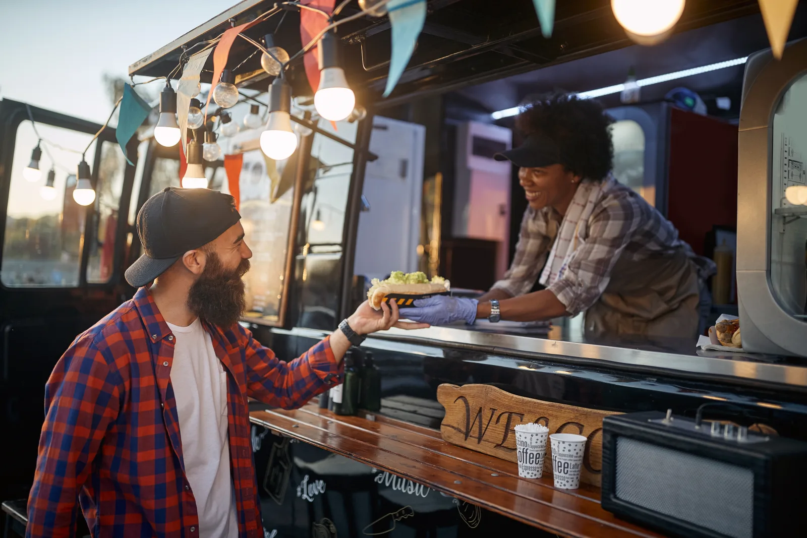 a wide view of a service hatch at golden hour, an order passing across the counter, takeaway cups and a wooden sign on the ledge — food truck insurance in Massachusetts