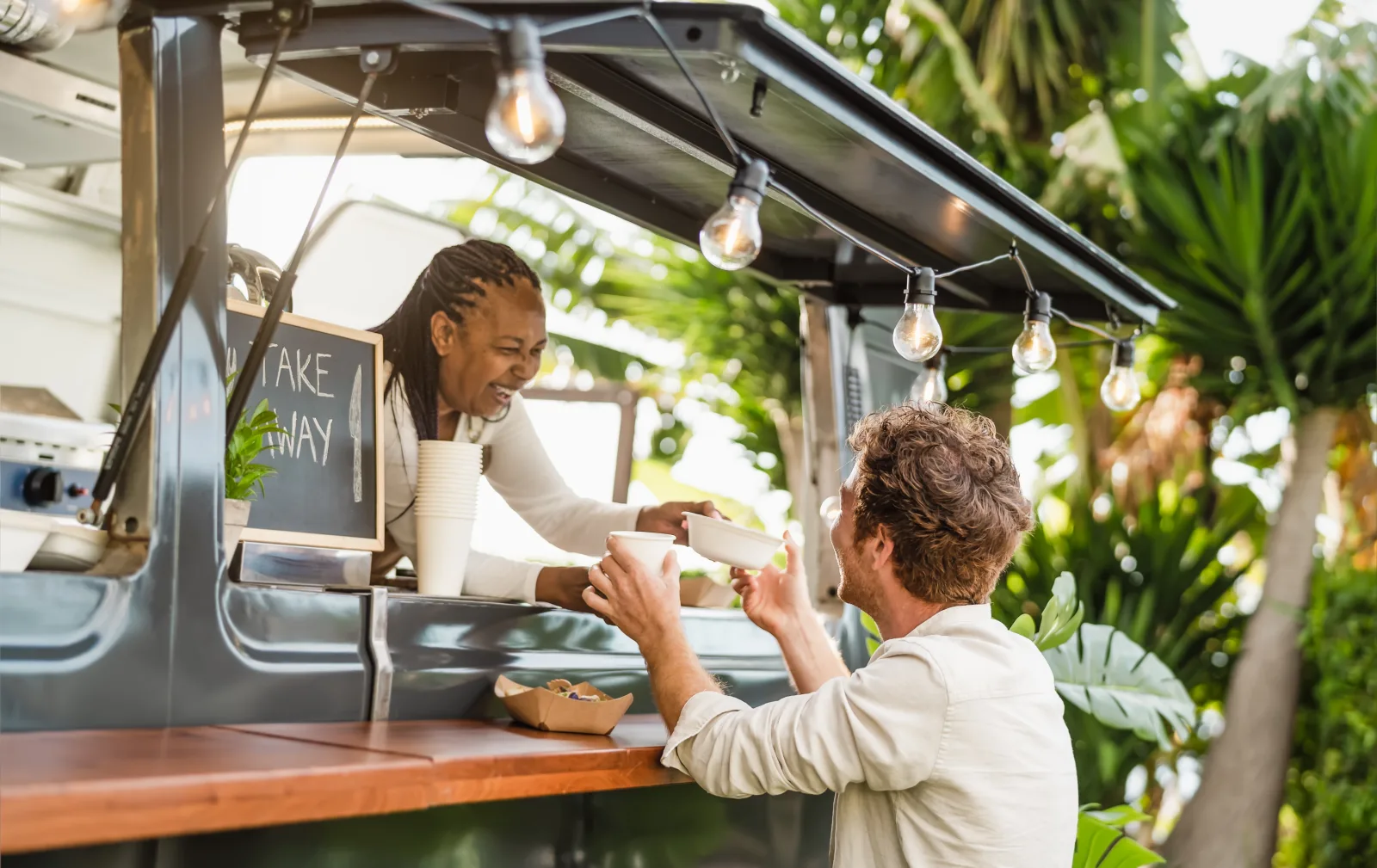 a gray food truck with a chalkboard propped in the window, a staff member handing a bowl across the counter, dense tropical planting behind — owner resources: are food trucks profitable? the real numbers for 2026
