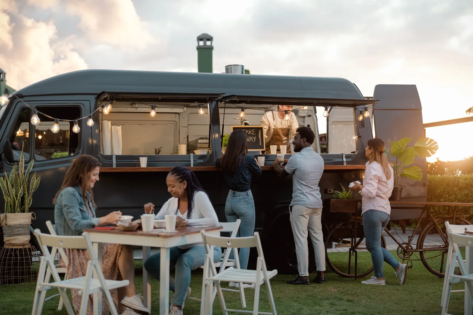 a dark van-bodied food truck on grass at sunset with a small queue at the hatch and customers eating at folding tables — food truck insurance in Wisconsin