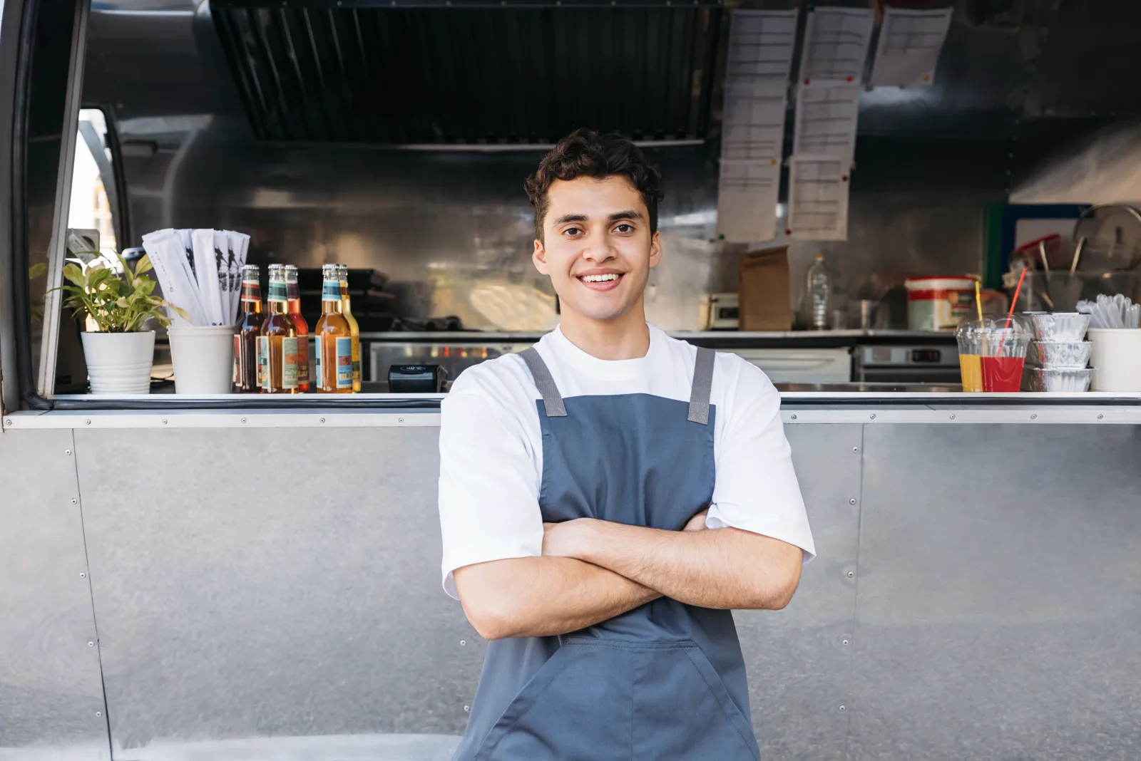 a staff member in an apron standing with arms folded in front of a brushed steel truck flank, bottles and ticket rail visible through the hatch — food truck insurance in Georgia