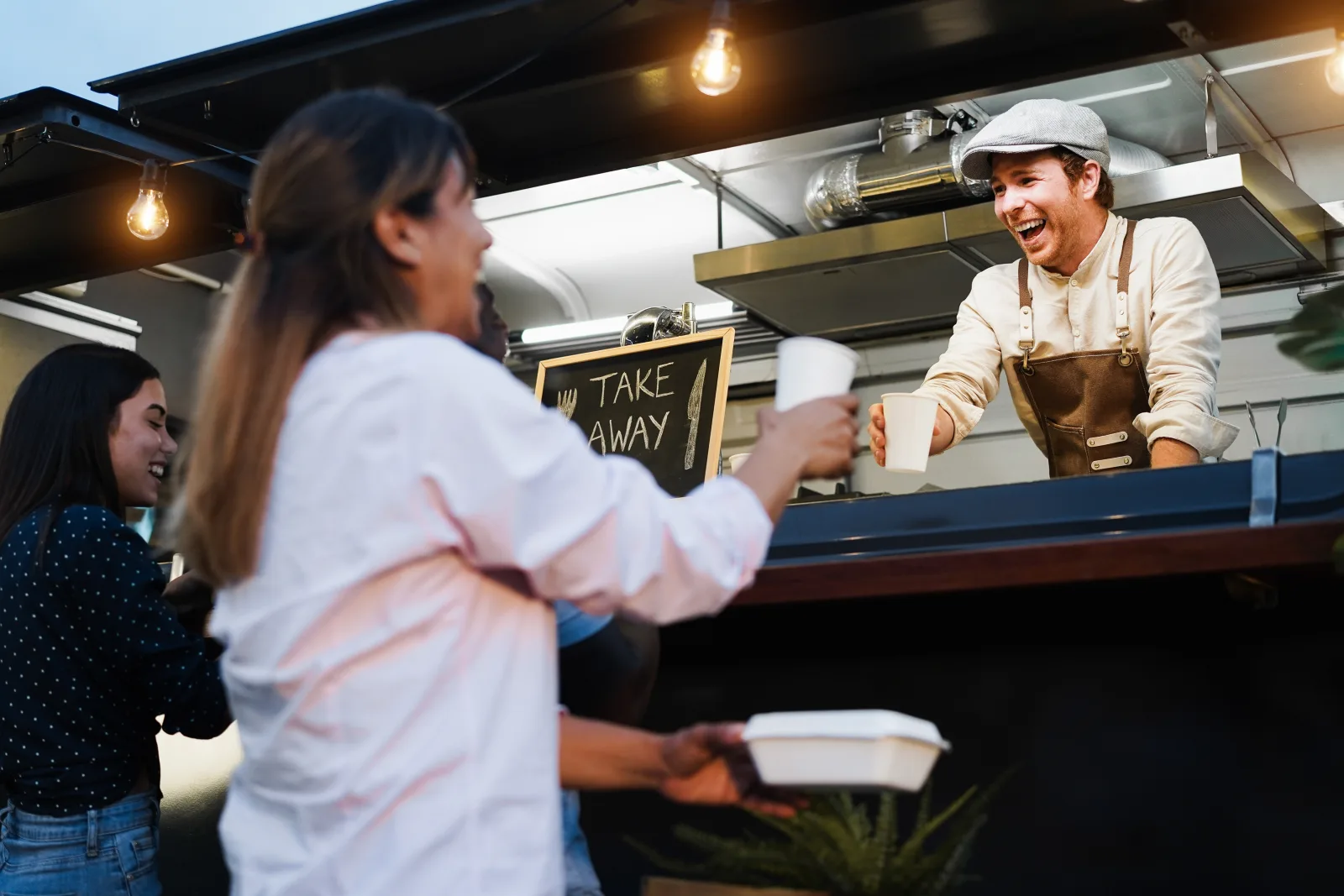 a staff member in a flat cap passing takeaway cups over a counter to two customers, a chalkboard sign beside the window — food truck insurance in Idaho
