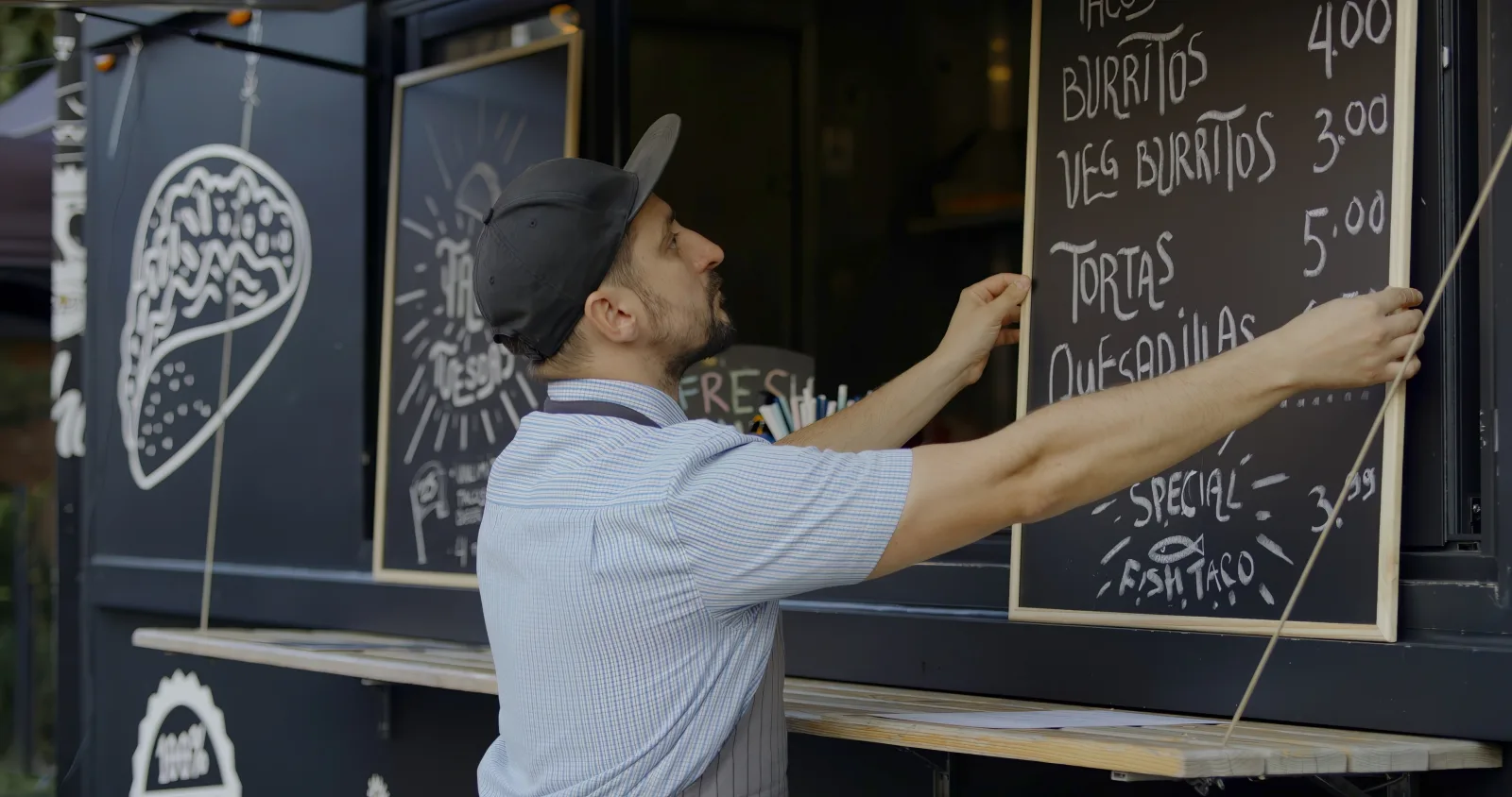 a staff member adjusting a framed chalkboard menu mounted on the side of a dark truck, a line-drawn taco painted on the panel beside it — food truck insurance in Illinois
