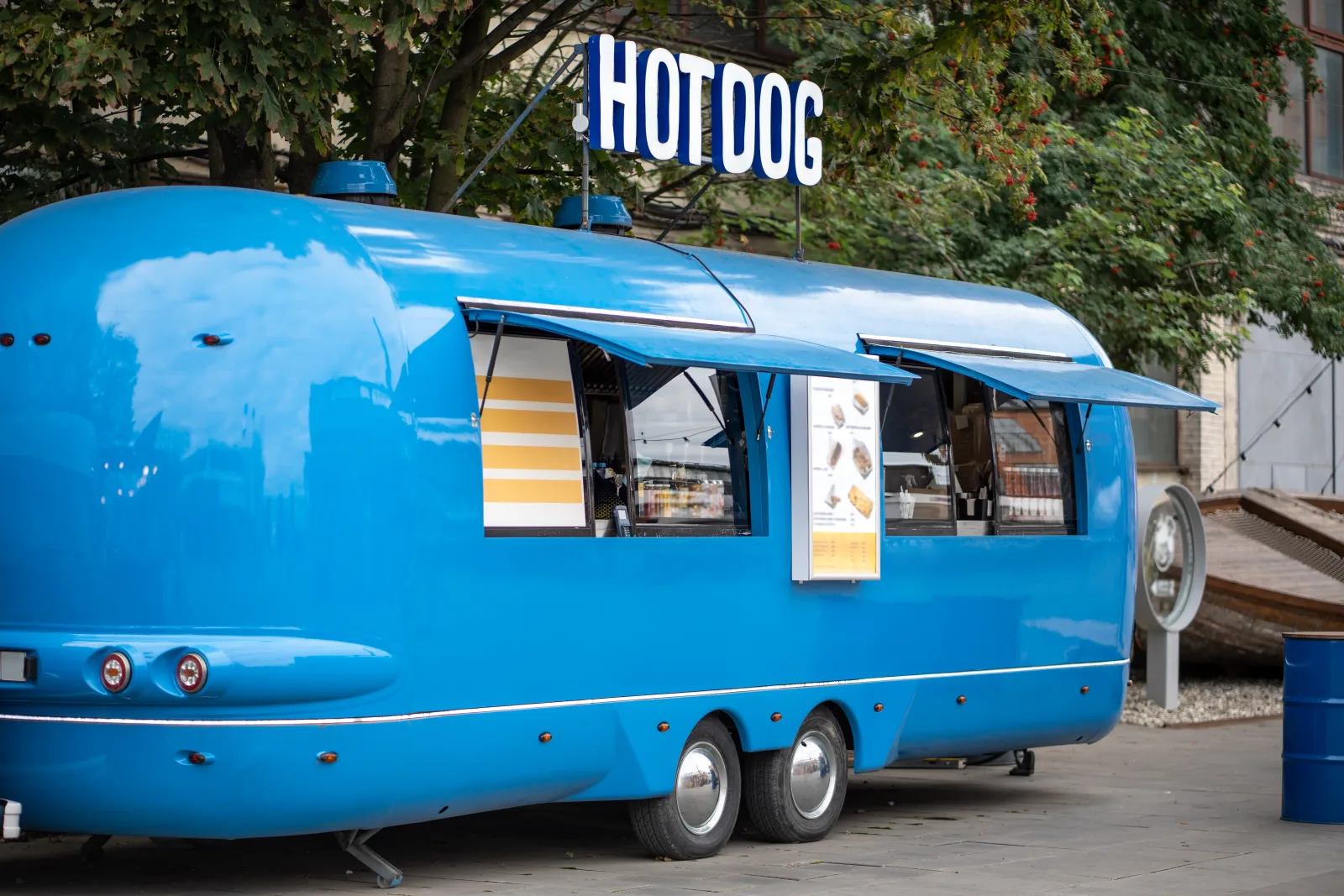 a bright blue riveted catering trailer with a raised sign board and two shuttered service windows, parked on paving under trees — food truck insurance in Indiana