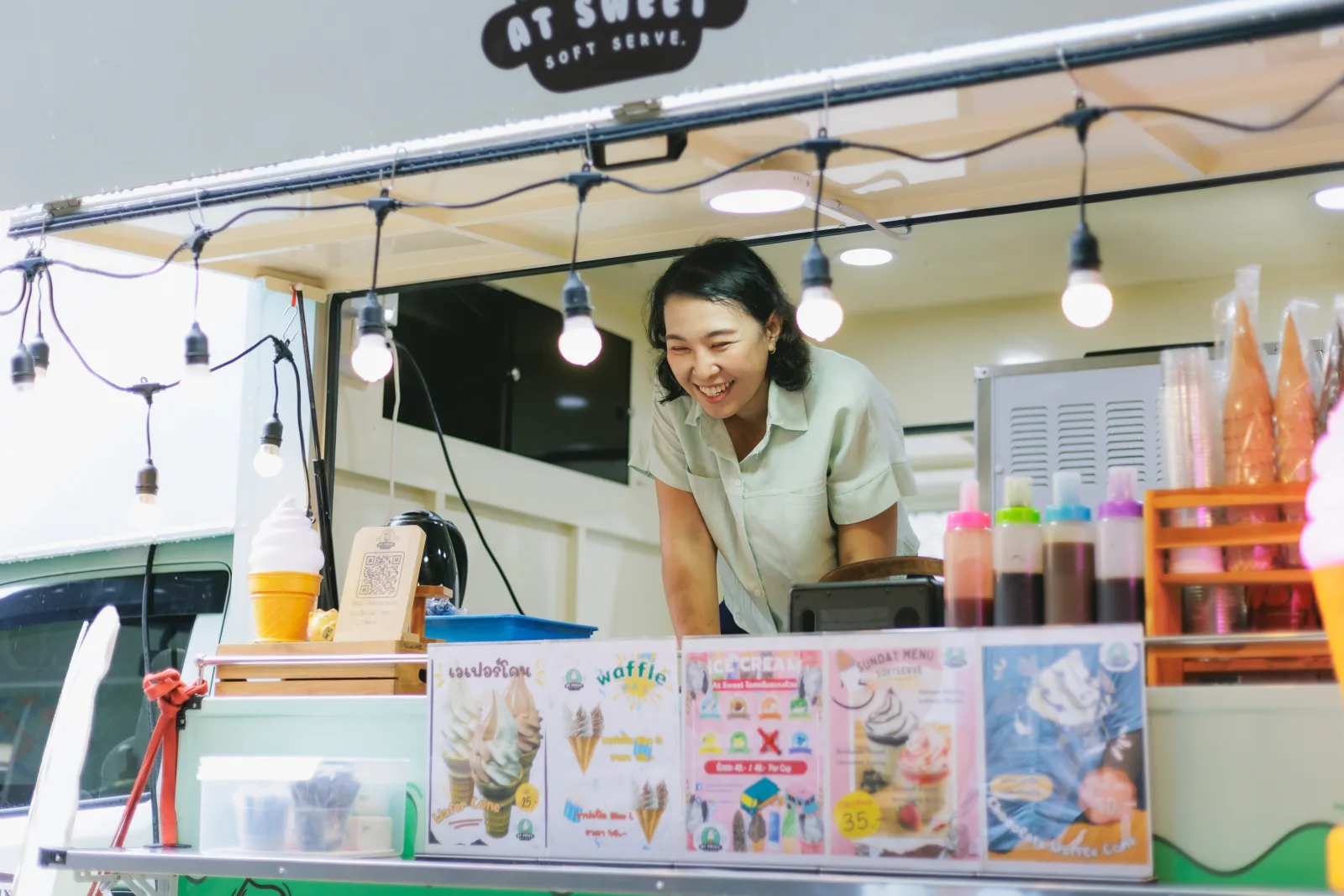 a smiling vendor leaning out of the service window of a soft-serve trailer, string lights along the awning, menu boards and sauce bottles ranged along the counter