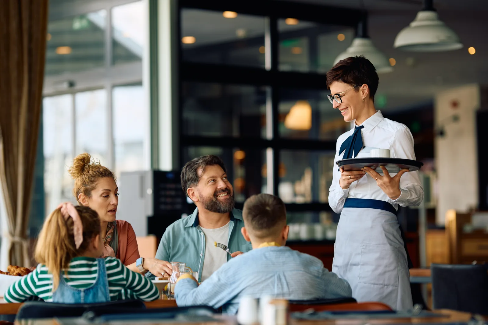 a server carrying a tray past a family seated at a window table — the agency behind Wexford Restaurant Insurance