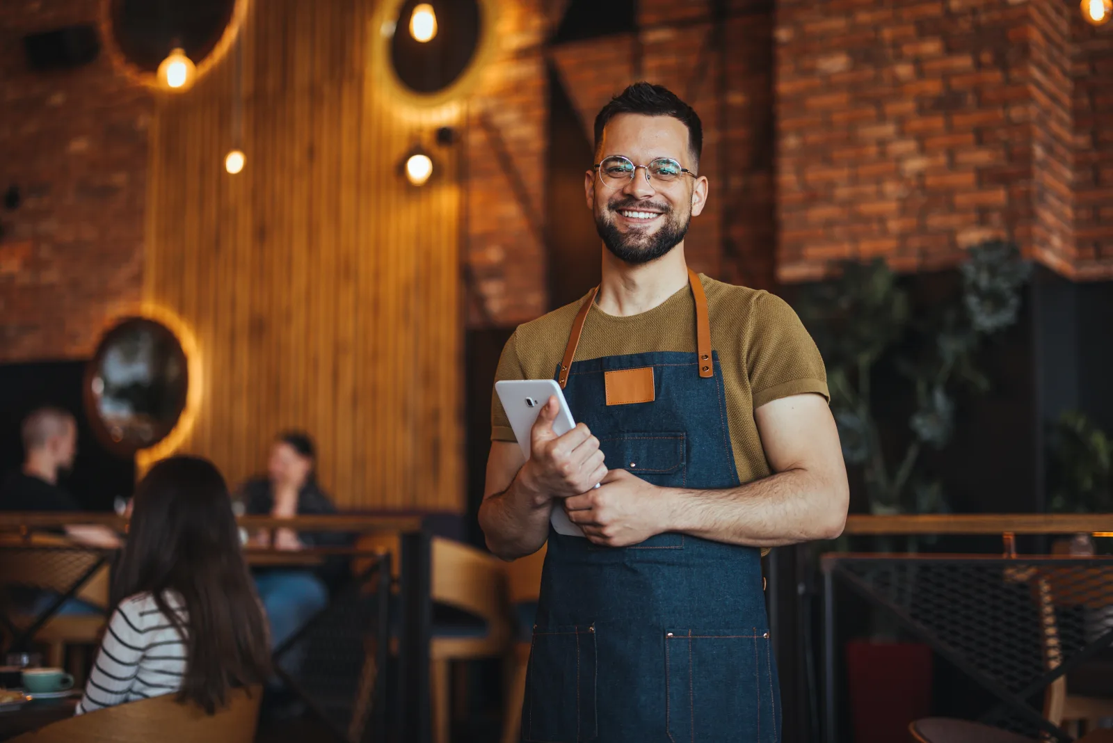 a man in a denim apron standing in a dining room holding a tablet — restaurant insurance in Georgia