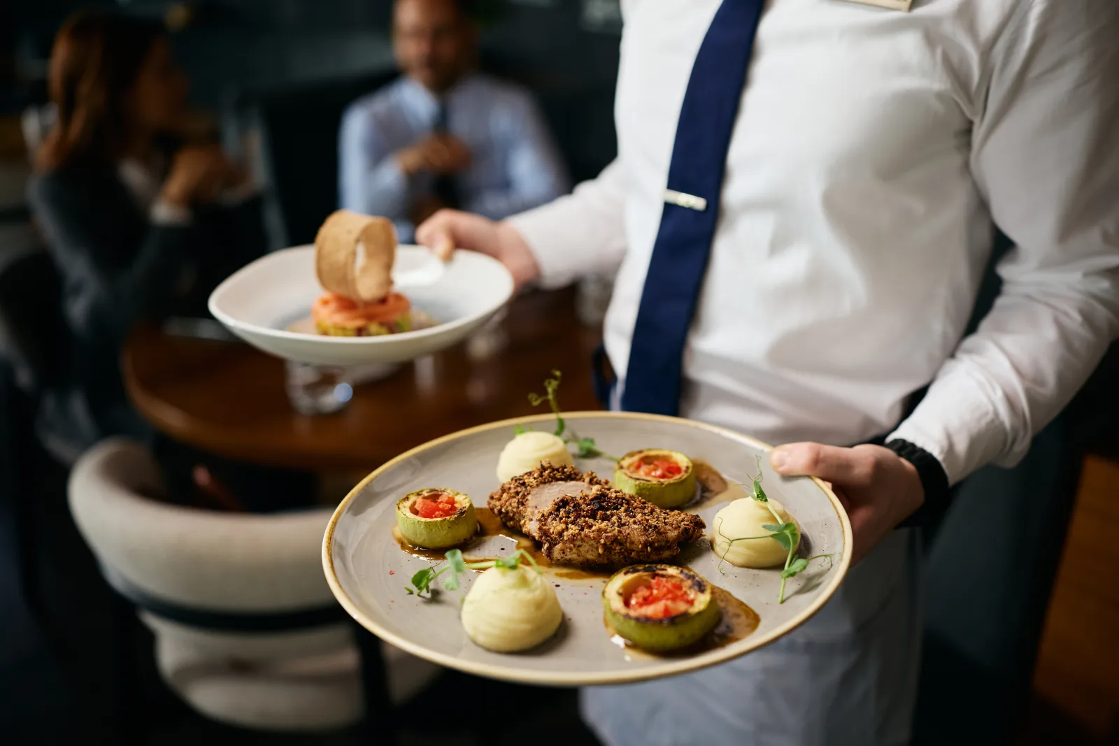 a server in a white shirt and tie carrying two plated dishes — restaurant insurance in North Carolina